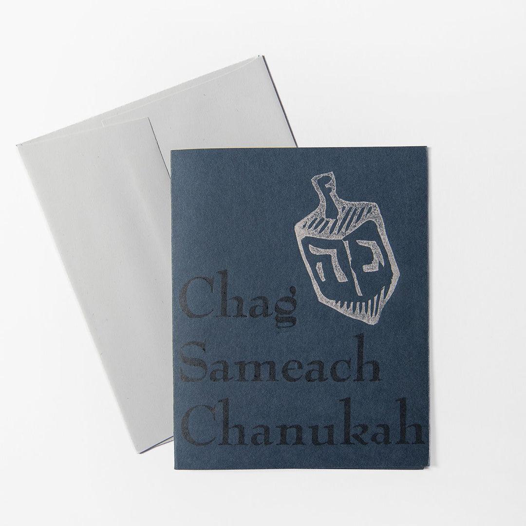 The Hanukkah Card features a blue design with a silver dreidel and bold "Chag Sameach Chanukah" text. Hand-printed by Liberty Craftworks, it comes with a white envelope, all set against a crisp white background.