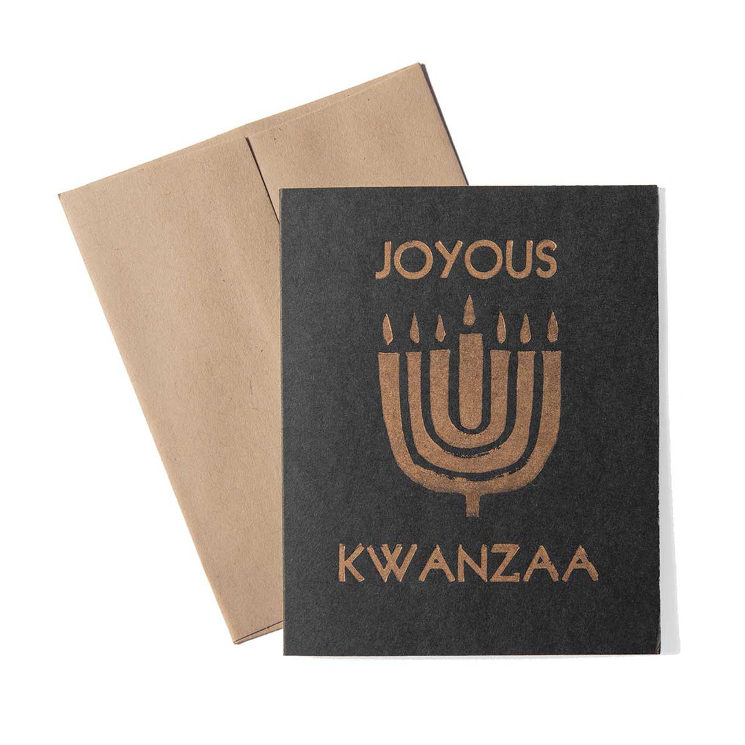 The Kwanzaa Card, crafted from black paper with a gold menorah and "Joyous Kwanzaa" message, is displayed next to a brown envelope on a white background.