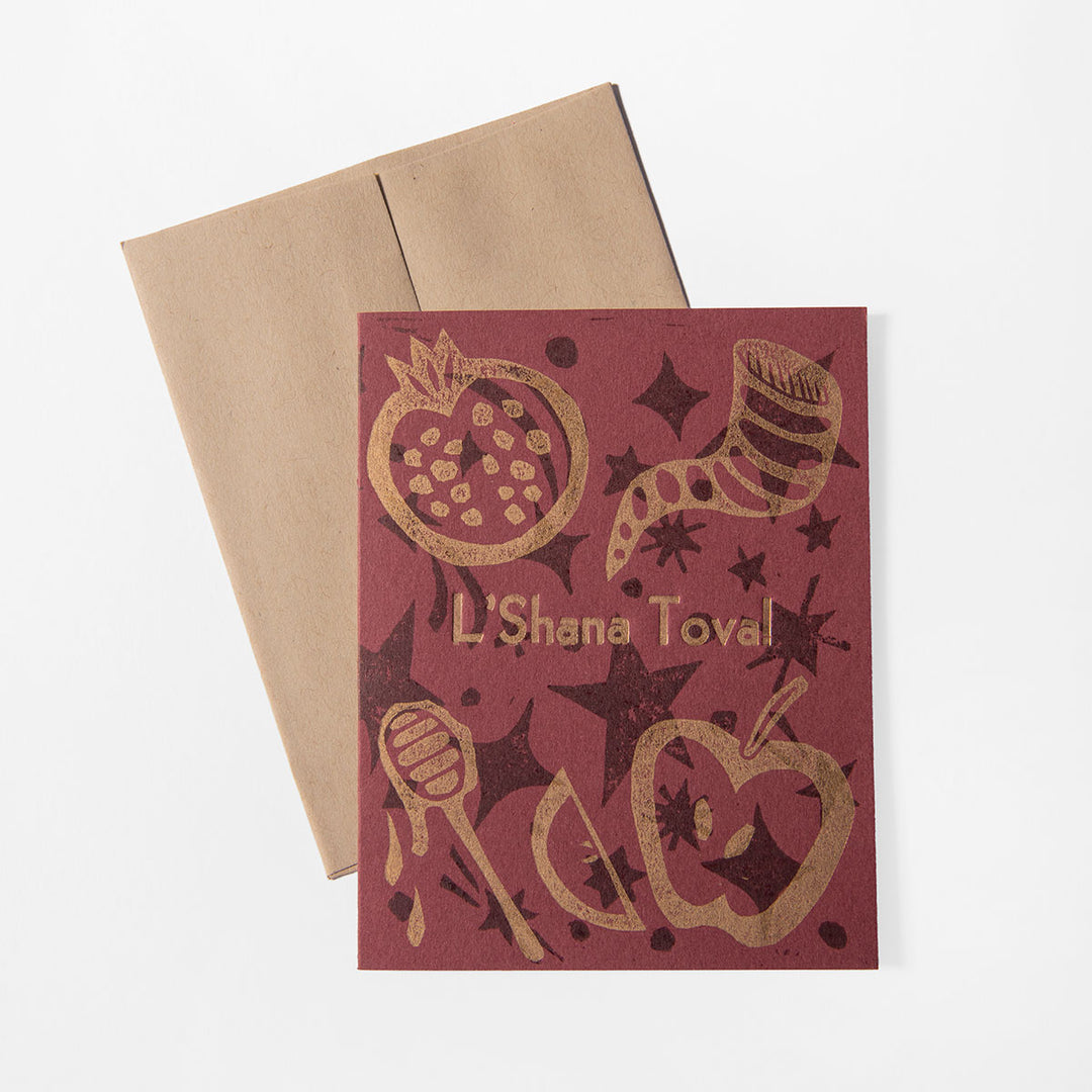 A maroon Rosh Hashanah Card by Liberty Craftworks, hand-printed with gold pomegranate, apple, honey dipper, shofar, and stars. “L’Shana Tova!” appears on the front. Includes a brown envelope behind the card.