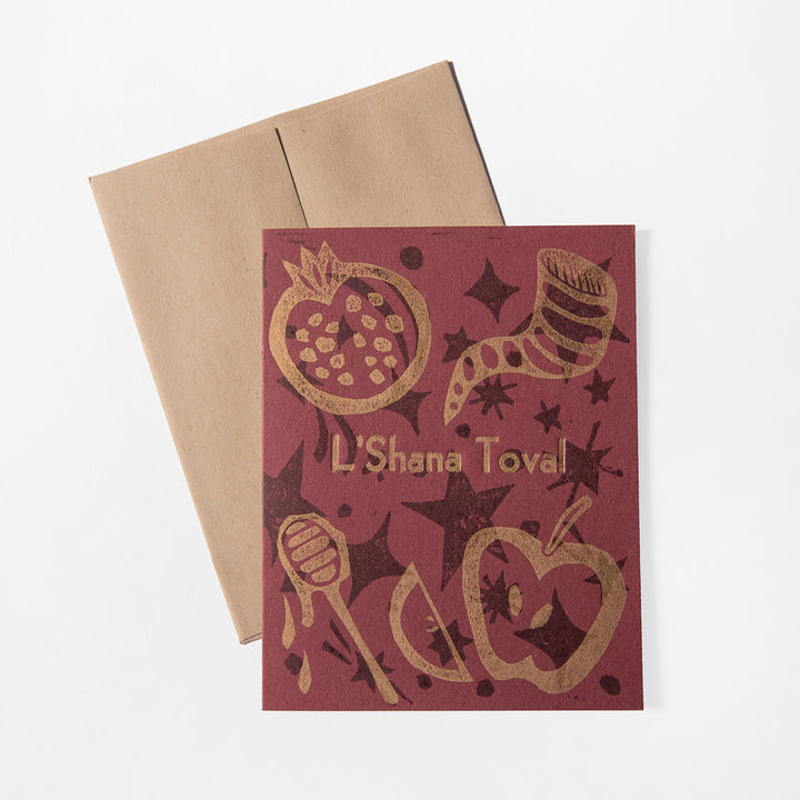 A maroon Rosh Hashanah Card by Liberty Craftworks, hand-printed with gold pomegranate, apple, honey dipper, shofar, and stars. “L’Shana Tova!” appears on the front. Includes a brown envelope behind the card.