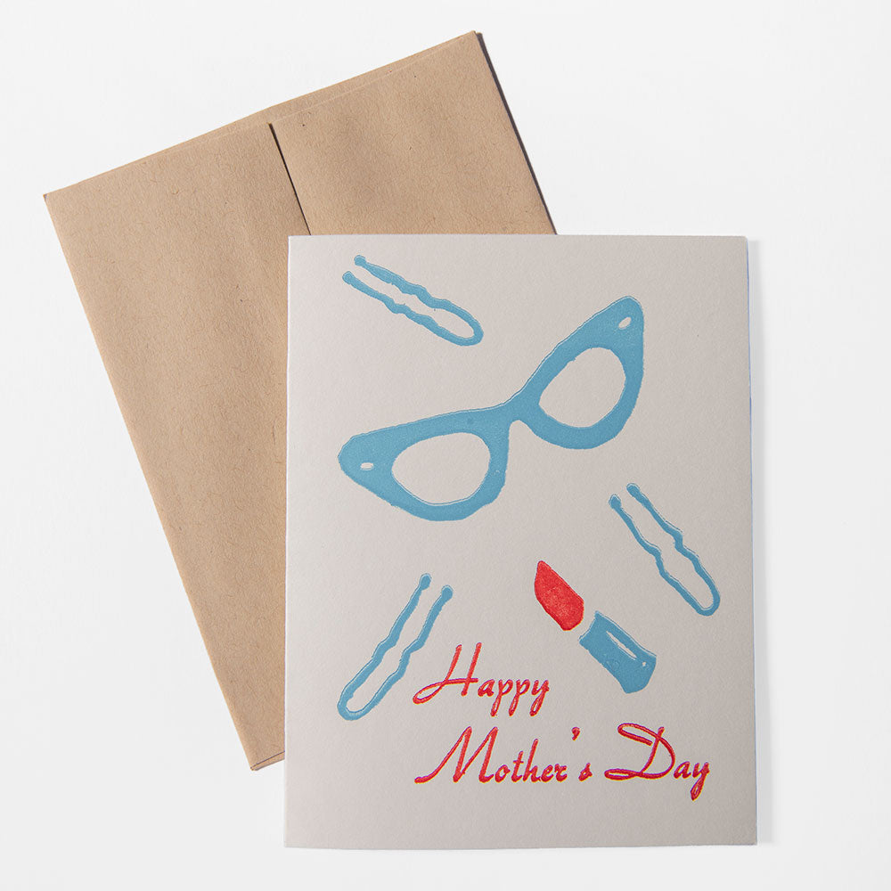 The Mother's Day Card by Liberty Craftworks is handcrafted with blue eyeglasses, hairpins, red lipstick, and “Happy Mother’s Day” in red script—paired with a brown envelope.
