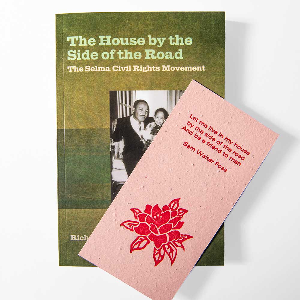The House by the Side of the Road Bookmark is displayed on a white background, overlapping with a pink card featuring a red rose illustration and an inspirational excerpt from a Sam Walter Foss poem, alongside the book "The Selma Civil Rights Movement." The bookmark is handcrafted from seeded paper.