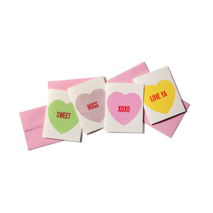 The Conversation Heart Card Set includes four small cards with pastel heart designs and messages—SWEET, HUGS, XOXO, LOVE YA—plus matching pink envelopes for each. Perfectly arranged on white, they're inspired by classic Valentine’s candy.