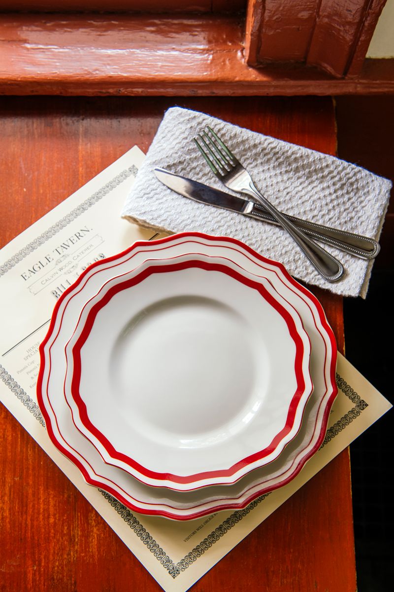 Three white plates with red rims are stacked on a menu, next to an America 250 Handwoven Napkin Set holding a fork and knife, all arranged on a wooden surface by a window.