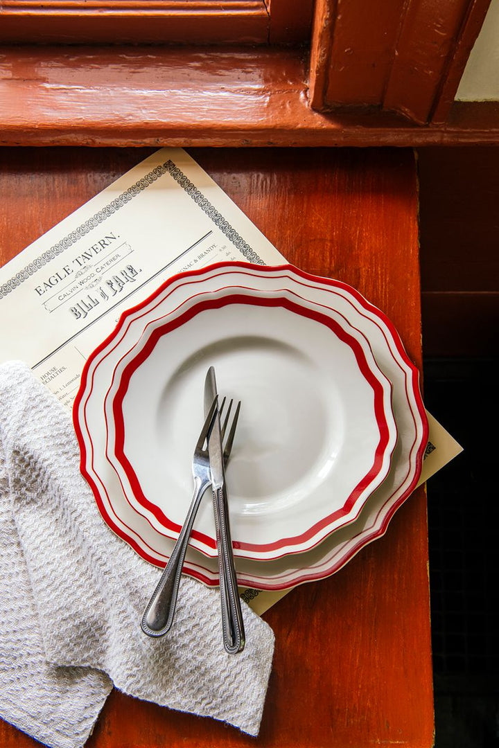 Sure! Here’s a revised version featuring the product name: Two white plates with red rims are stacked on a wooden table, topped with a crossed fork and knife, while an America 250 Handwoven Napkin Set—crafted in the tradition of Greenfield Village weaving—adds texture alongside a partially visible menu in the background.