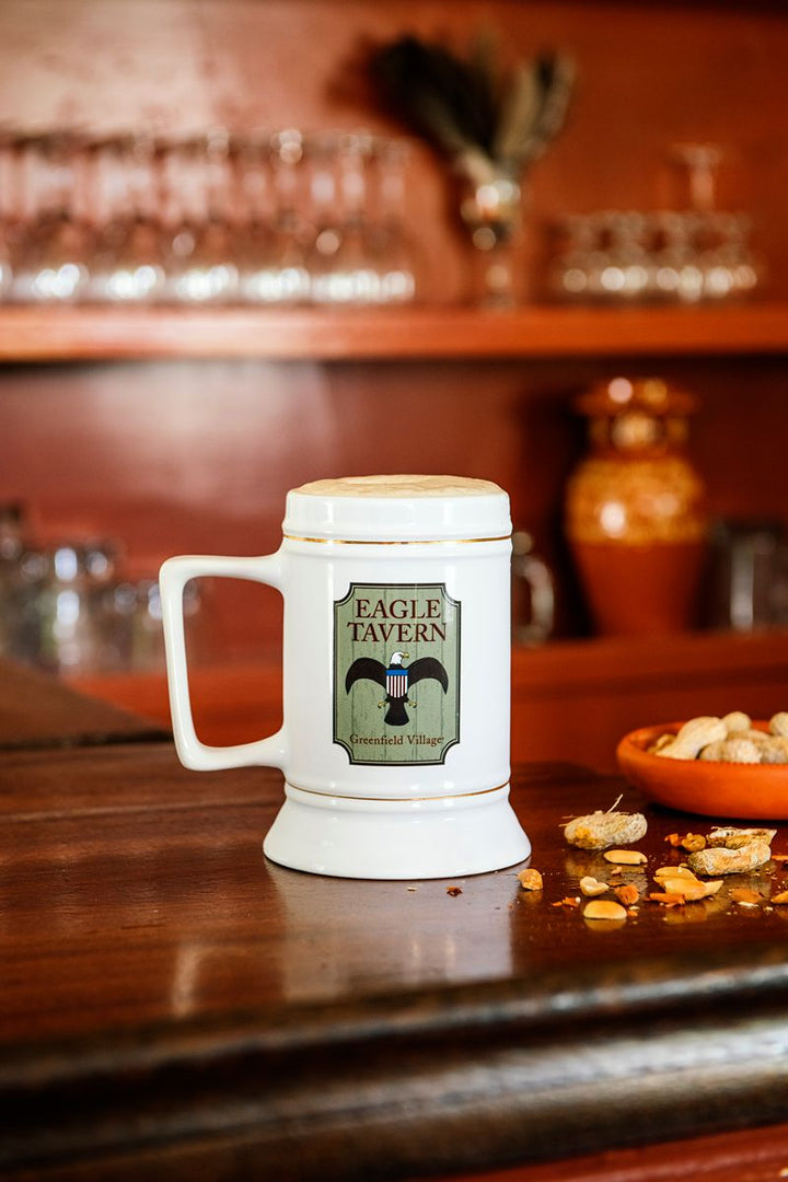 A Greenfield Village Eagle Tavern Stein, emblazoned with “Eagle Tavern,” rests on a wooden bar beside a bowl of peanuts, conjuring the nostalgic charm of 19th-century America. In the background, shelves filled with glassware and pottery evoke the historic ambiance of Greenfield Village.