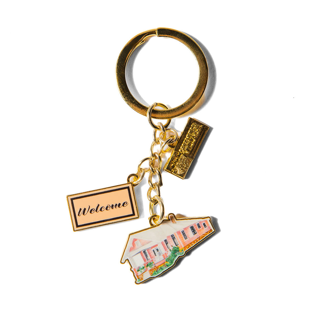 Gold keychain featuring three charms: a rectangular Welcome sign, a horizontal bar, and a small enamel house-shaped charm with a pink roof and front porch, all displayed on a white background. The Greenfield Village Jackson Home Dangle Keychain makes an ideal Greenfield Village souvenir or thoughtful gift.