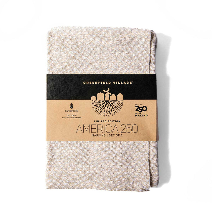 The America 250 Handwoven Napkin Set includes two beige, textured cloth napkins made with traditional Greenfield Village weaving techniques. The napkins are wrapped in a brown paper label featuring “GREENFIELD VILLAGE AMERICA 250,” along with “Handwoven Cotton” and “Limited Edition.” This set elegantly blends historical craftsmanship with enduring style.