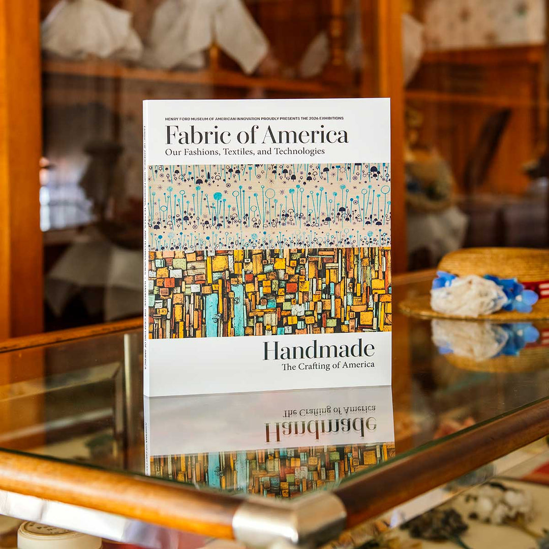 The Henry Ford’s Fabric of America and Handmade Exhibition Catalog stands on a glass display case with textiles and hats in the background, offering a glimpse into American artifacts showcased at the Fabric of America exhibition.
