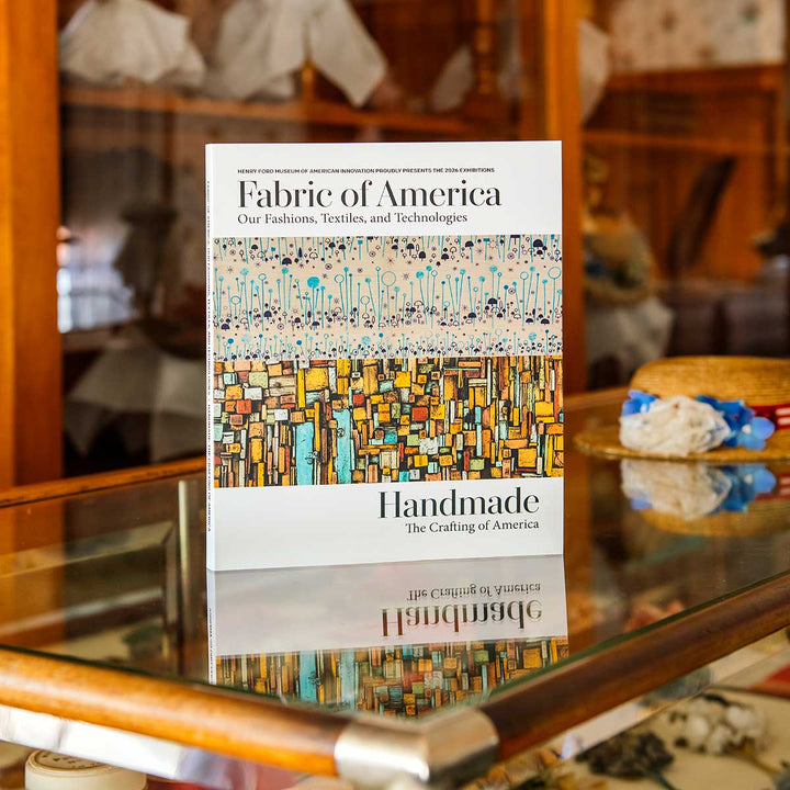 The Henry Ford’s Fabric of America and Handmade Exhibition Catalog stands on a glass display case with textiles and hats in the background, offering a glimpse into American artifacts showcased at the Fabric of America exhibition.