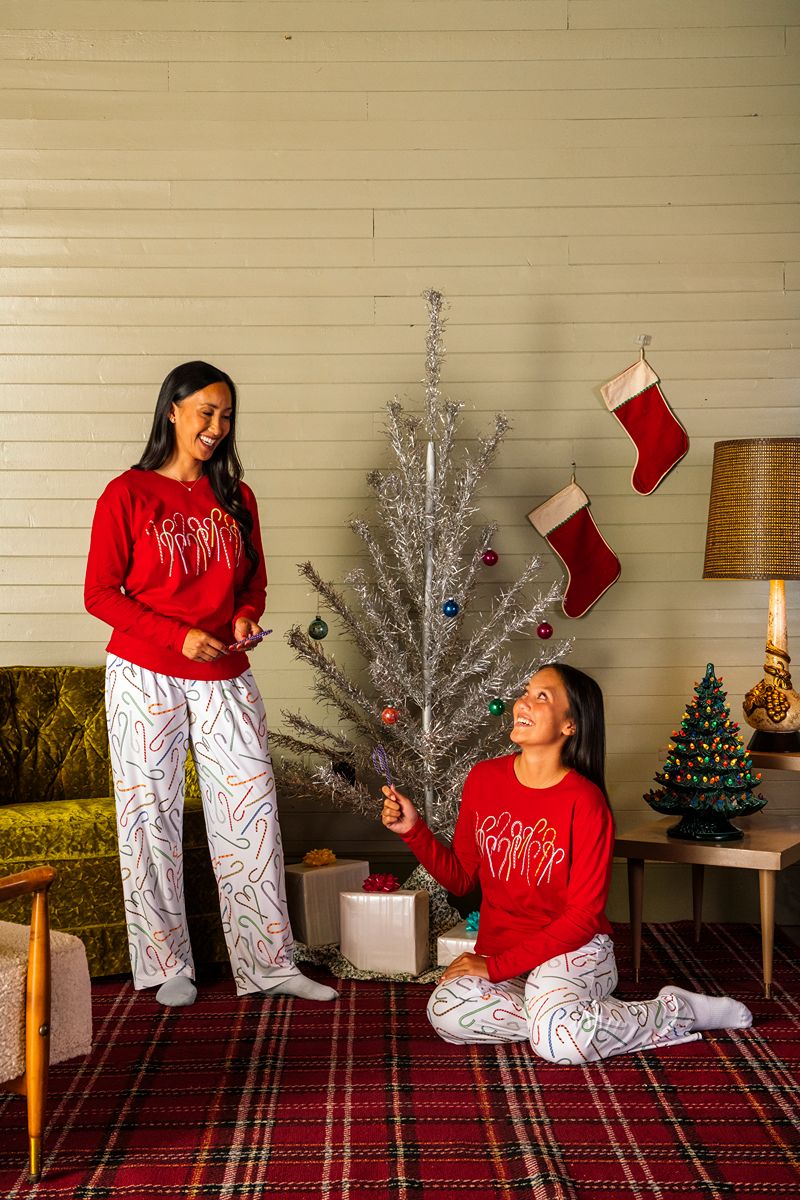 Two women in matching holiday pajamas decorate a silver Christmas tree in a cozy living room adorned with stockings, wrapped gifts, a vintage lamp, and a ceramic Christmas tree on a plaid carpet. One of them is wearing The Henry Ford® Greenfield Village Candy Cane Pajama Shirt - Red, adding extra cheer to the warm holiday setting.