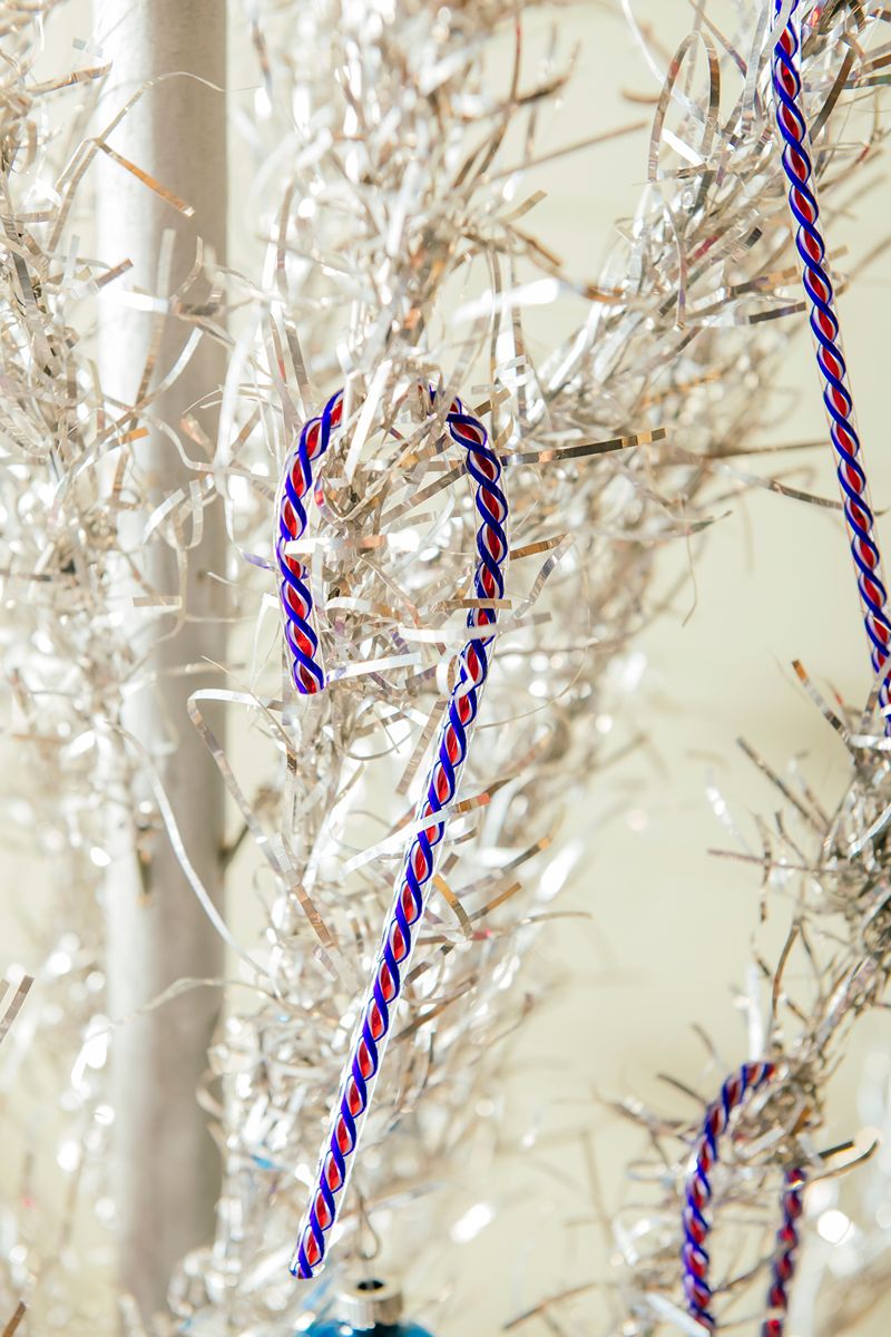 A shiny silver tinsel Christmas tree is adorned with the 2026 Candy Cane—a limited-edition red, white, and blue ornament specially crafted for America’s 250th—hanging from its sparkling branches.