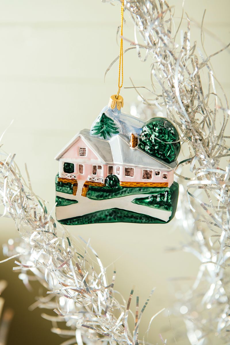 A ceramic house ornament featuring green trees and a yellow door—The Henry Ford® Jackson Home Ornament—hangs on a silver tinsel Christmas tree, with sunlight illuminating the scene and evoking a charming, historic keepsake.