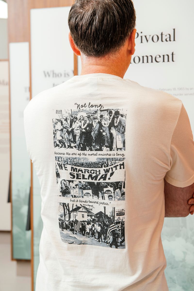 A man stands with arms crossed, wearing the Greenfield Village Selma How Long History T-Shirt, which features black-and-white images and text about a civil rights march. The T-shirt highlights a quote from Martin Luther King Jr. about justice and references the Selma March.