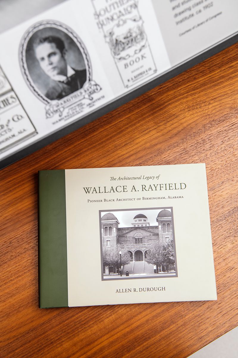 The book, The Architectural Legacy of Wallace A. Rayfield: Pioneer Black Architect of Birmingham, Alabama by Allen R. Durough, rests on a wooden surface; its cover displays a striking black-and-white photograph of a building that honors the work of trailblazing African American architect Wallace Rayfield, with a historical photo partially visible above.