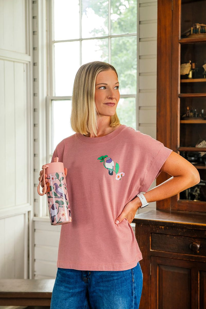 Wearing the Roberto Lugo Pretty Pigeon Women’s T-Shirt, which features a striking bird graphic, a woman stands indoors beside a window and wooden cabinet, holding a pink-patterned water bottle and gazing to the side with a relaxed demeanor. This art-inspired tee brings together modern style and artistic expression, making it an ideal choice for outings such as trips to the Henry Ford Museum.
