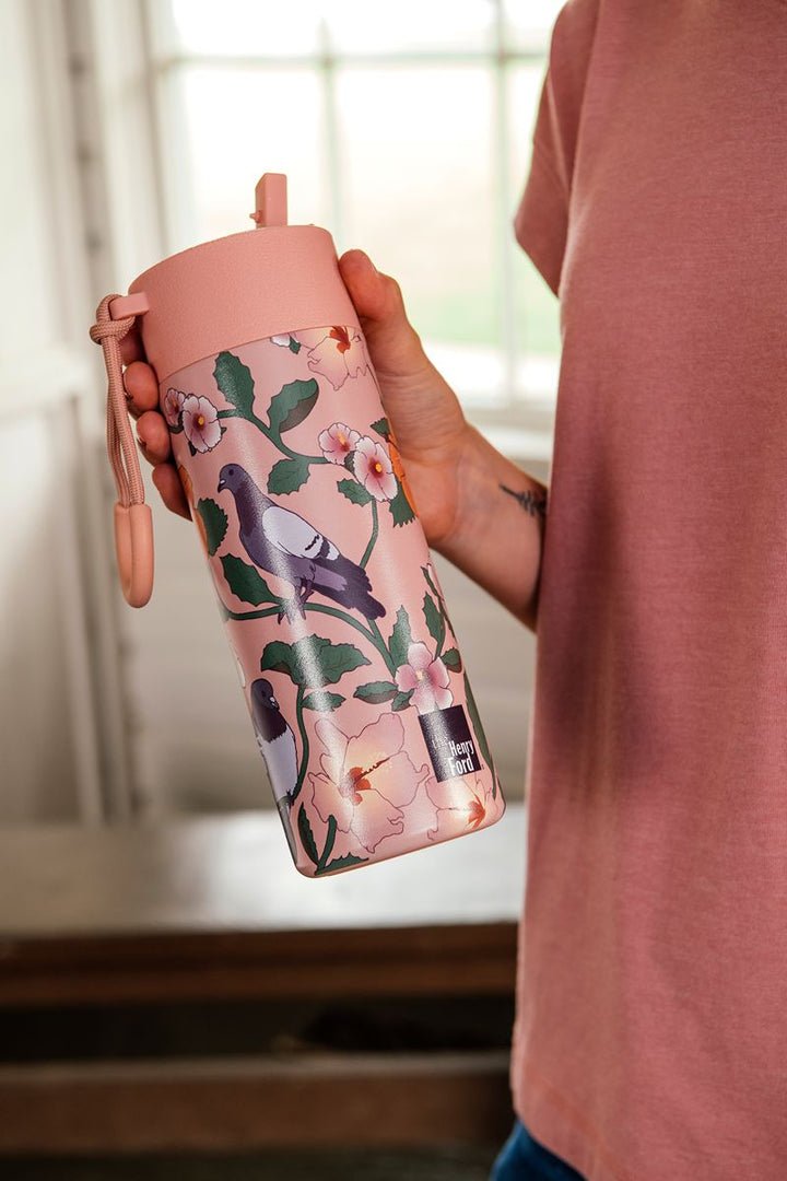 A person stands indoors near a window, dressed in a pink shirt and blue jeans, holding the Roberto Lugo Pigeon Art Water Bottle—a pink 32 oz insulated stainless steel bottle adorned with floral and bird designs.
