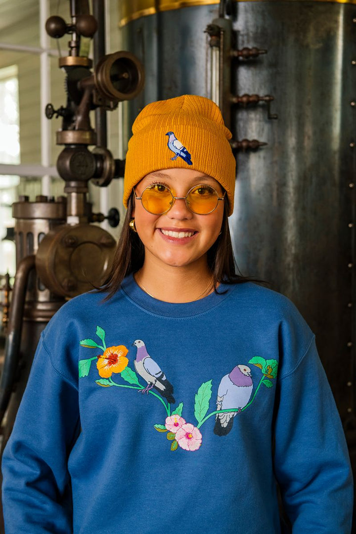 A smiling person wearing yellow glasses, a mustard beanie with a bird design, and the Roberto Lugo Pigeon Crew Sweatshirt—a blue museum gift sweatshirt featuring bird and flower embroidery—stands in front of vintage industrial machinery.