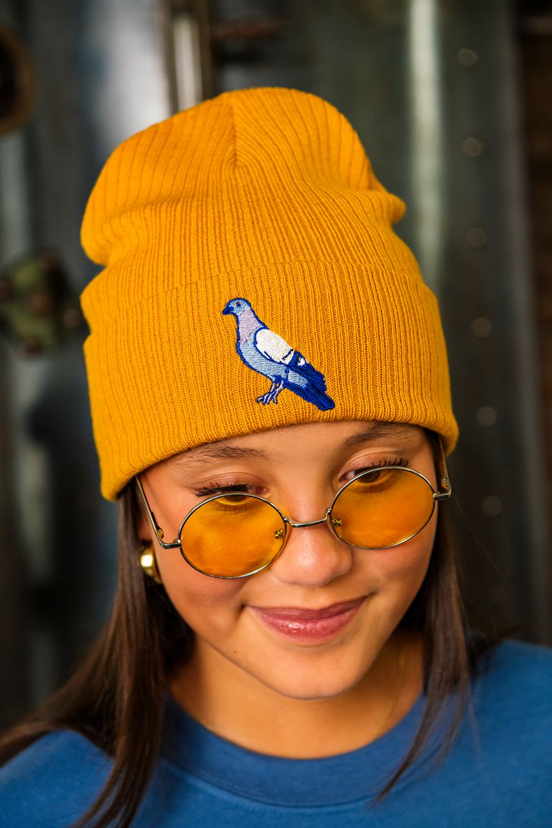 A person smiles gently while wearing round yellow-tinted glasses and the Roberto Lugo Pigeon Beanie—mustard yellow and embroidered with a blue and white pigeon—embodying an artful heritage inspired by the creative spirit of The Henry Ford Museum.