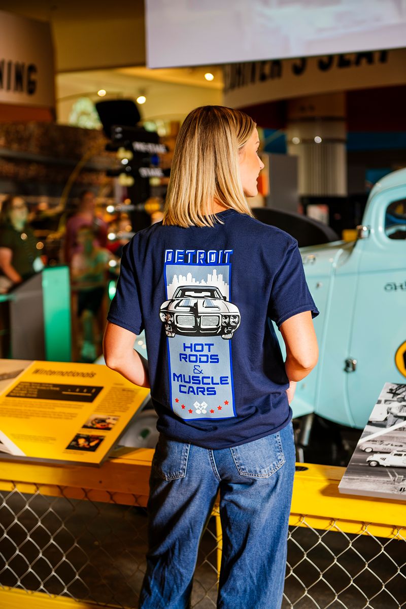 A woman with blonde hair, wearing The Henry Ford® Hot Rod Movie T-Shirt in navy and jeans, stands in front of a vintage car display at a museum.