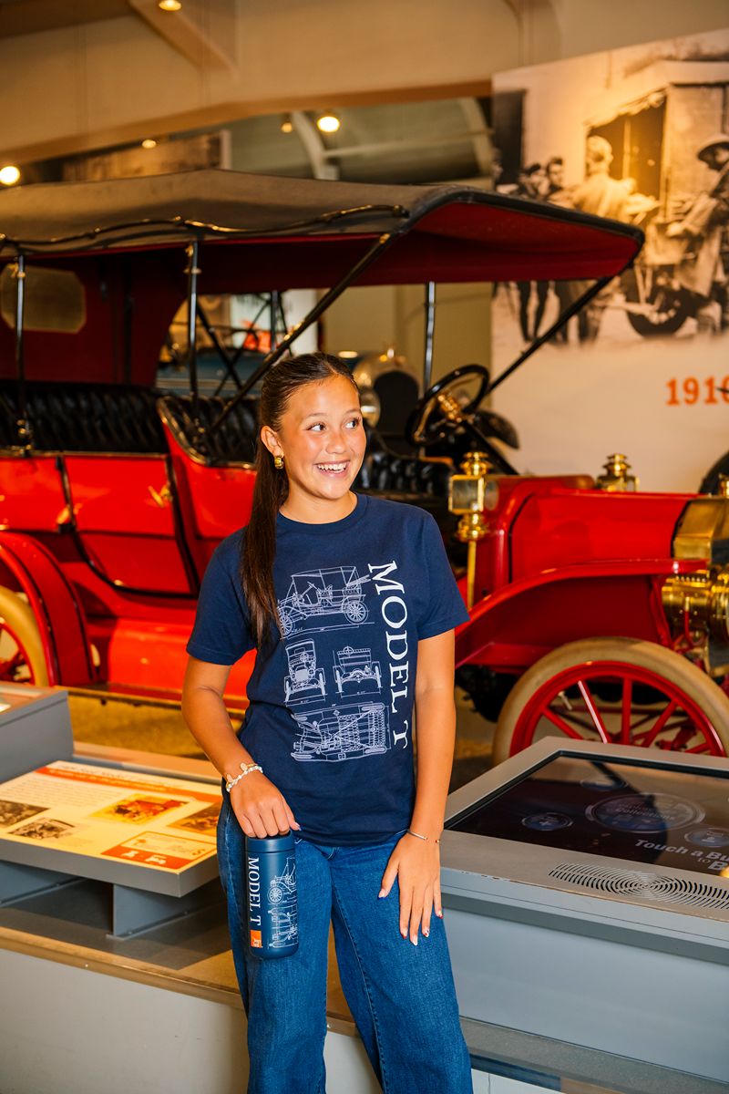 A smiling girl in The Henry Ford® Model T Tee Shirt stands in front of a red vintage car at a museum exhibit, holding a water bottle—perfectly capturing the spirit of the Henry Ford Museum.
