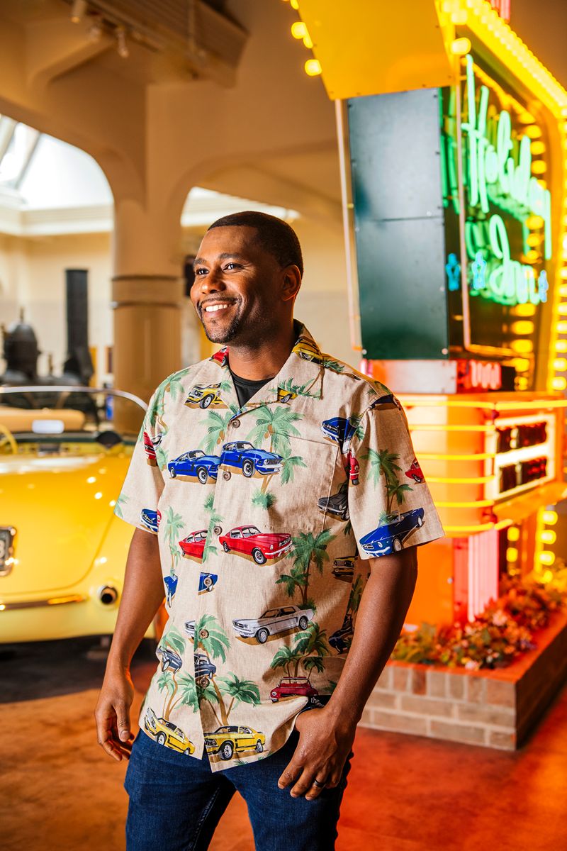 A man smiles while standing indoors near a neon sign and a yellow vintage car. He is wearing the Ford Old Mustangs Camp Shirt, featuring a colorful pattern of cars and palm trees. The setting appears retro with warm, bright lighting.