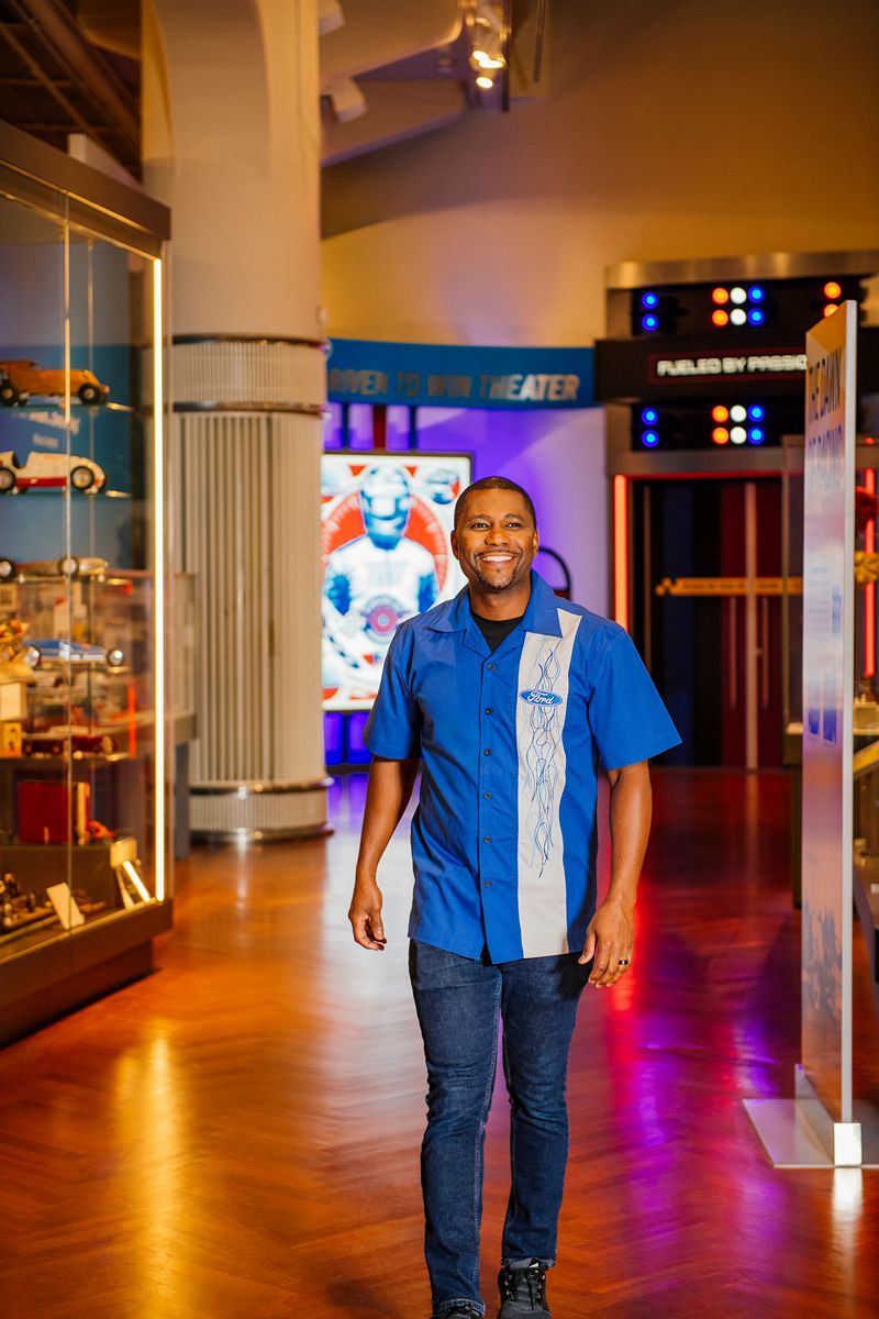 Wearing a Ford Flames Club Shirt, a man smiles as he strolls through a brightly lit museum filled with classic American automobiles, vibrant displays, and a Captain America poster—a perfect destination for car enthusiasts.