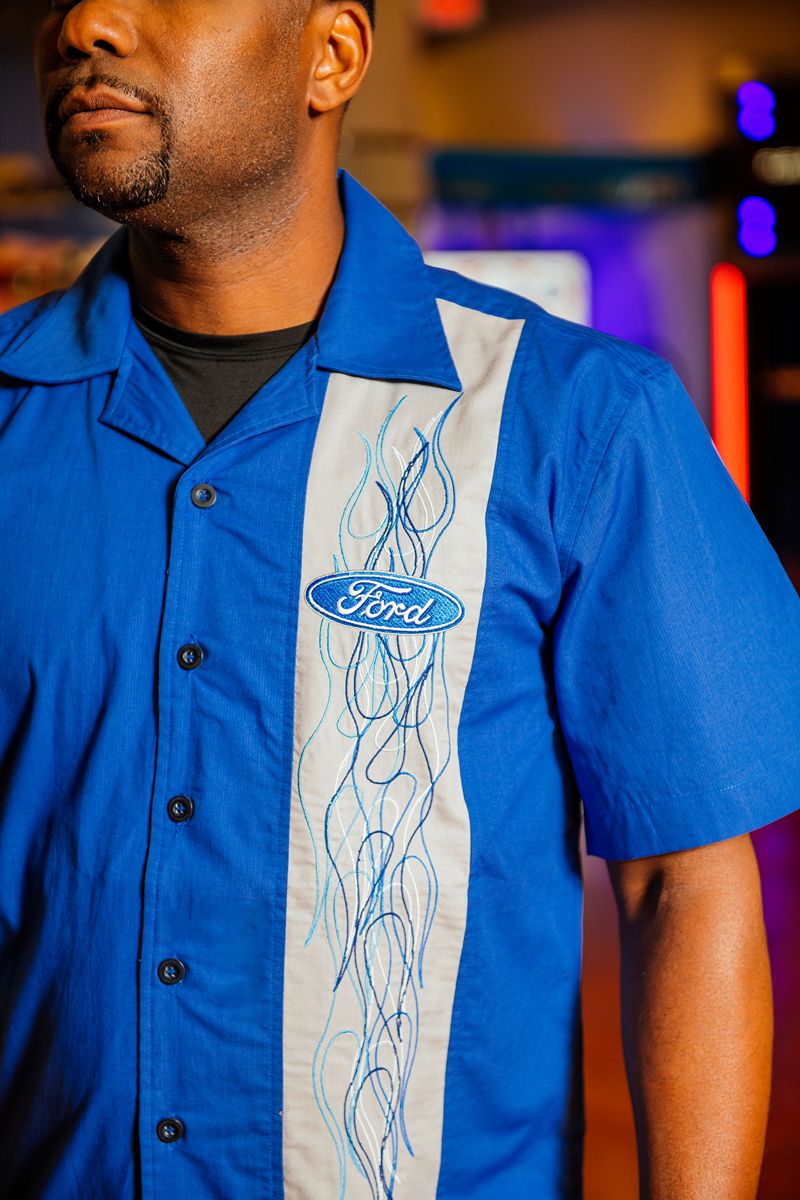 A man is seen indoors wearing the Ford Flames Club Shirt—a blue short-sleeve shirt with a white panel adorned with blue decorative lines and a Ford logo. Perfect for car enthusiasts, only his upper body and part of his face are visible.