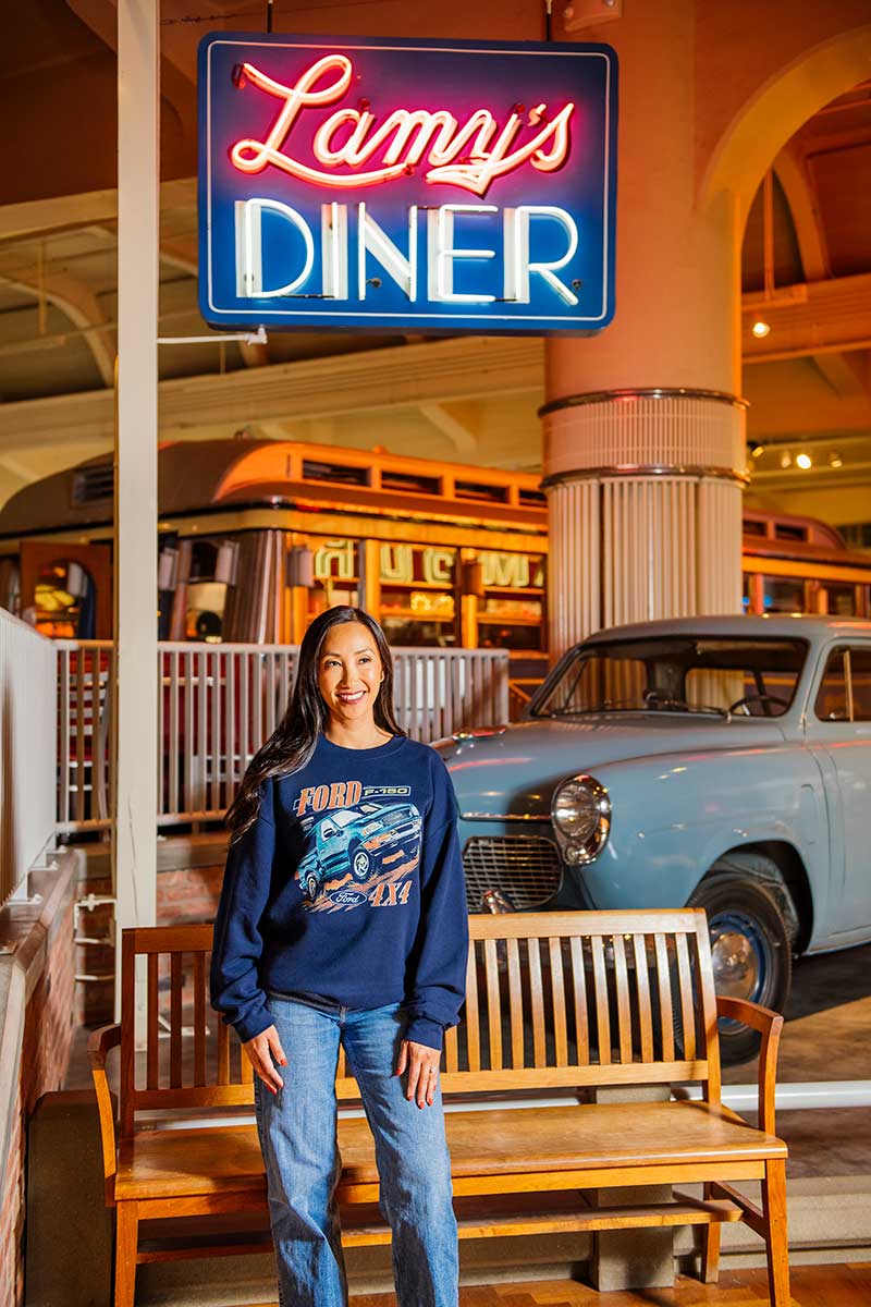 A woman wearing a Ford F-150 4x4 Crew Sweatshirt stands smiling beside a wooden bench under a glowing neon Lamys Diner sign, with a vintage blue car and the diner in the background—capturing the rugged durability that defines every Ford classic.