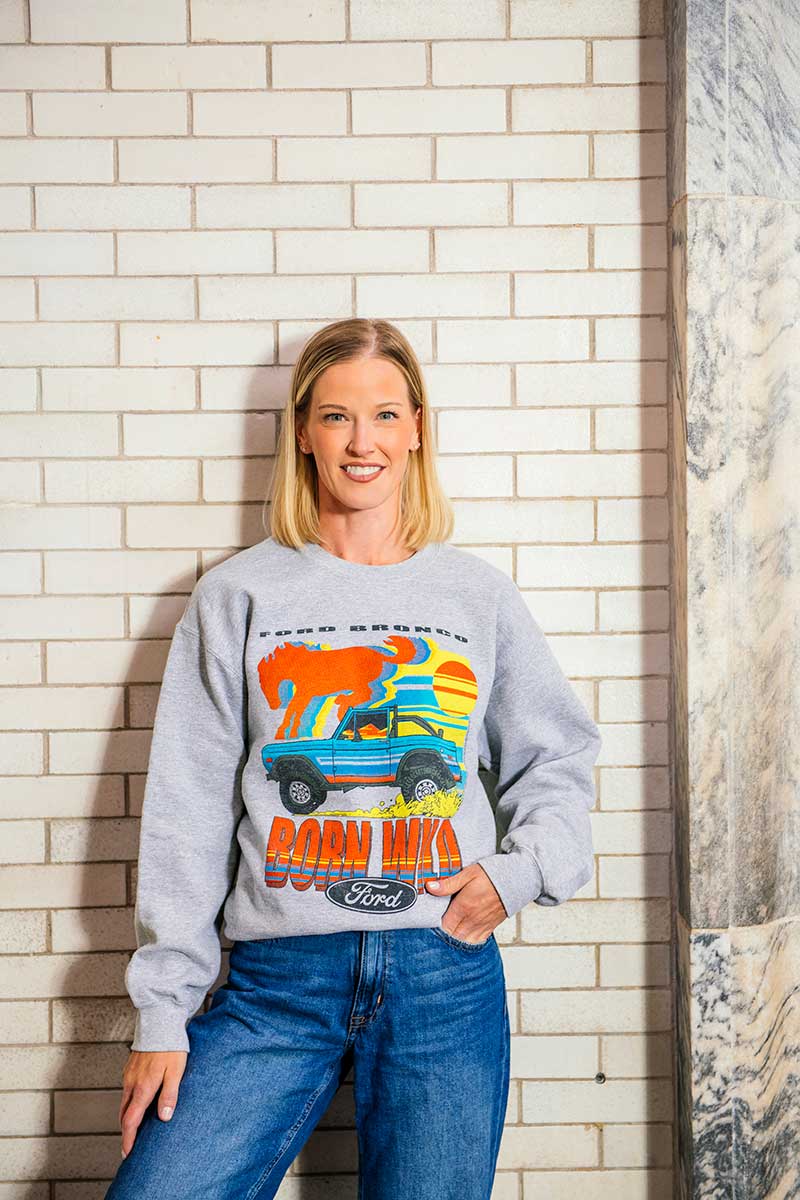 A woman with blond hair, dressed in blue jeans and the Ford Bronco Born Wild Fleece Sweatshirt, stands against a white brick wall. The sweatshirt showcases a vibrant graphic of a car, sun, and “Born Wild” text, perfectly capturing the adventurous spirit of the apparel.