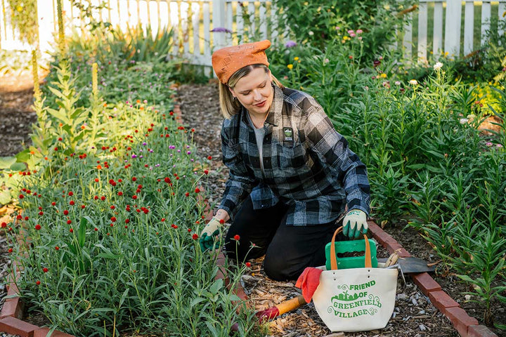 A woman wearing a plaid shirt and orange hat kneels in a garden, tending to red flowers. She holds gardening tools, with a Friend of Greenfield Village Project Tote and watering can beside her. Lush plants and a white fence are in the background.