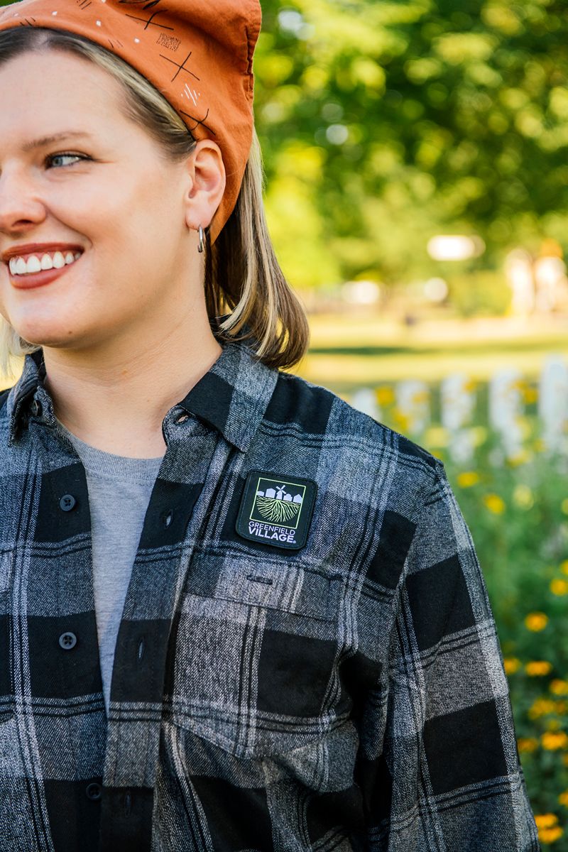 A smiling person in a Greenfield Village Flannel T-Shirt and an orange headscarf stands outdoors near flowers. A patch on the shirt reads “Village.” The background is green and sunny.