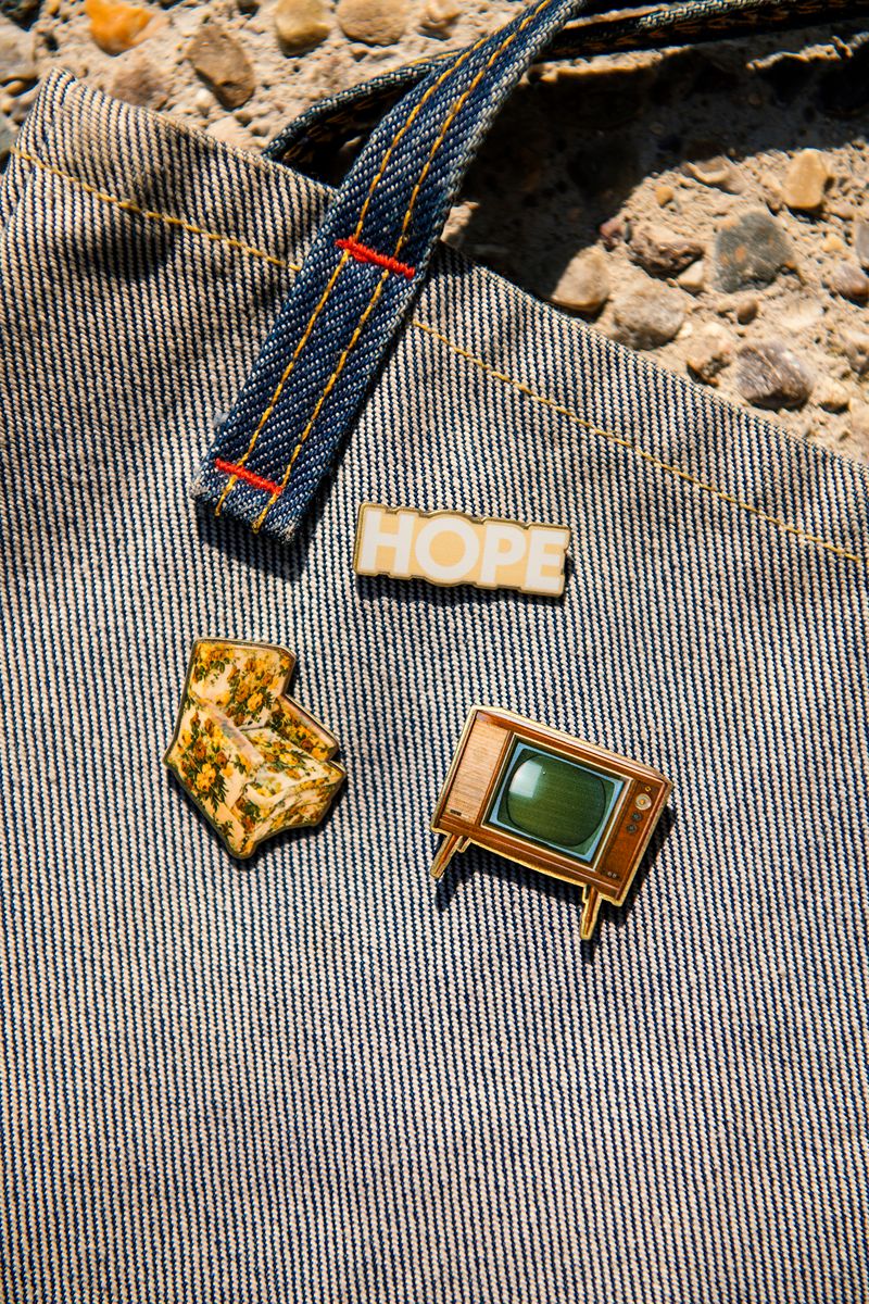 Three enamel pins on denim: a white HOPE pin, the Greenfield Village Jackson Home Retro Pin Set featuring a gold state-shaped Jackson Home pin, and a vintage television pin with a green screen. Stone ground background visible at the top, evoking the spirit of Greenfield Village.