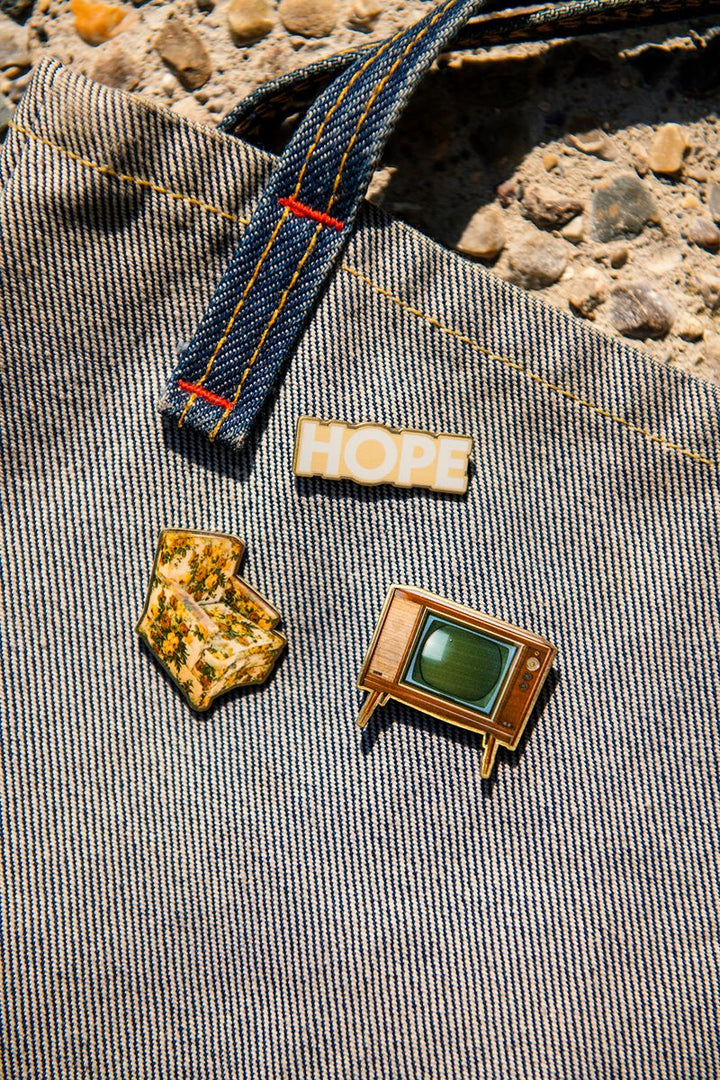 Three enamel pins on denim: a white HOPE pin, the Greenfield Village Jackson Home Retro Pin Set featuring a gold state-shaped Jackson Home pin, and a vintage television pin with a green screen. Stone ground background visible at the top, evoking the spirit of Greenfield Village.