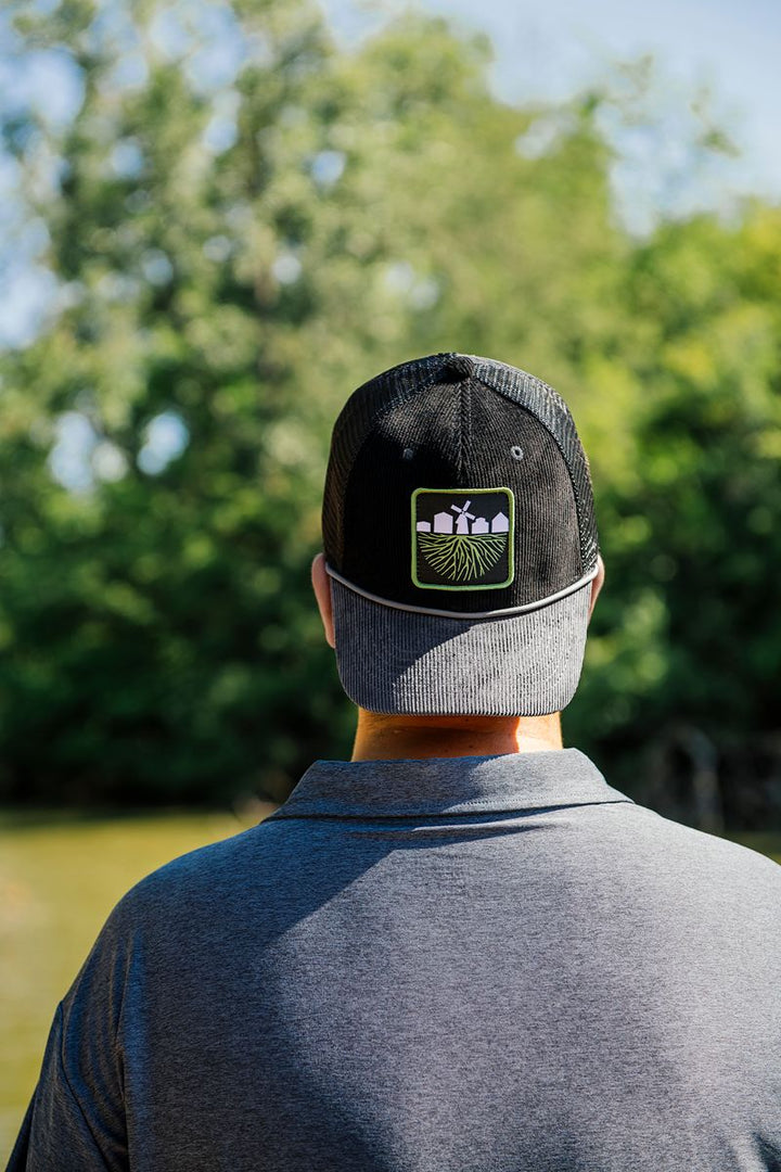 A person wearing a black mesh Greenfield Village Corduroy Trucker Cap with a farming logo stands outdoors facing away, trees and water softly blurred in the background—a perfect living history museum souvenir.
