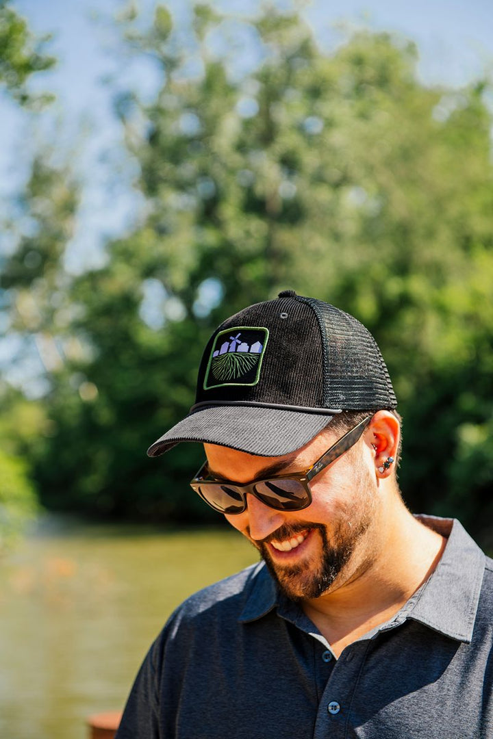 A man wearing sunglasses and a black hat styled like the Greenfield Village Corduroy Trucker Cap with a nature-themed patch smiles outdoors near water, with trees and a blue sky in the background.