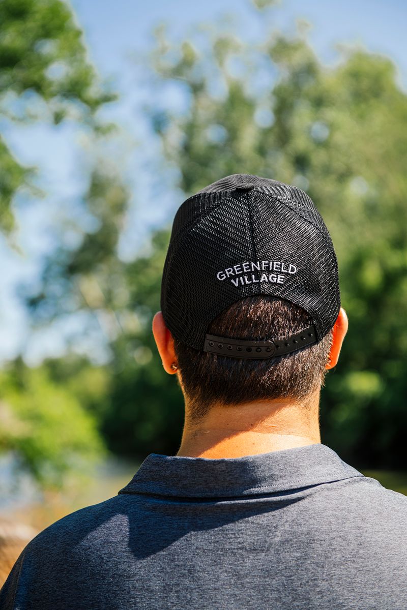 Wearing a black Greenfield Village Corduroy Trucker Cap—an iconic trucker cap style souvenir from the living history museum—a person stands outdoors with greenery and a blue sky in the background, seen from behind.