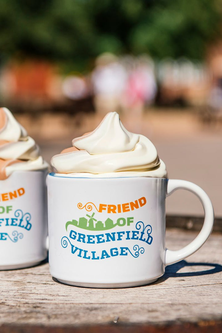 A Friend of Greenfield Village Mug filled with vanilla soft serve ice cream rests on a wooden surface outdoors. Next to it, another identical mug is partially visible, capturing the charm of this living history museum setting.