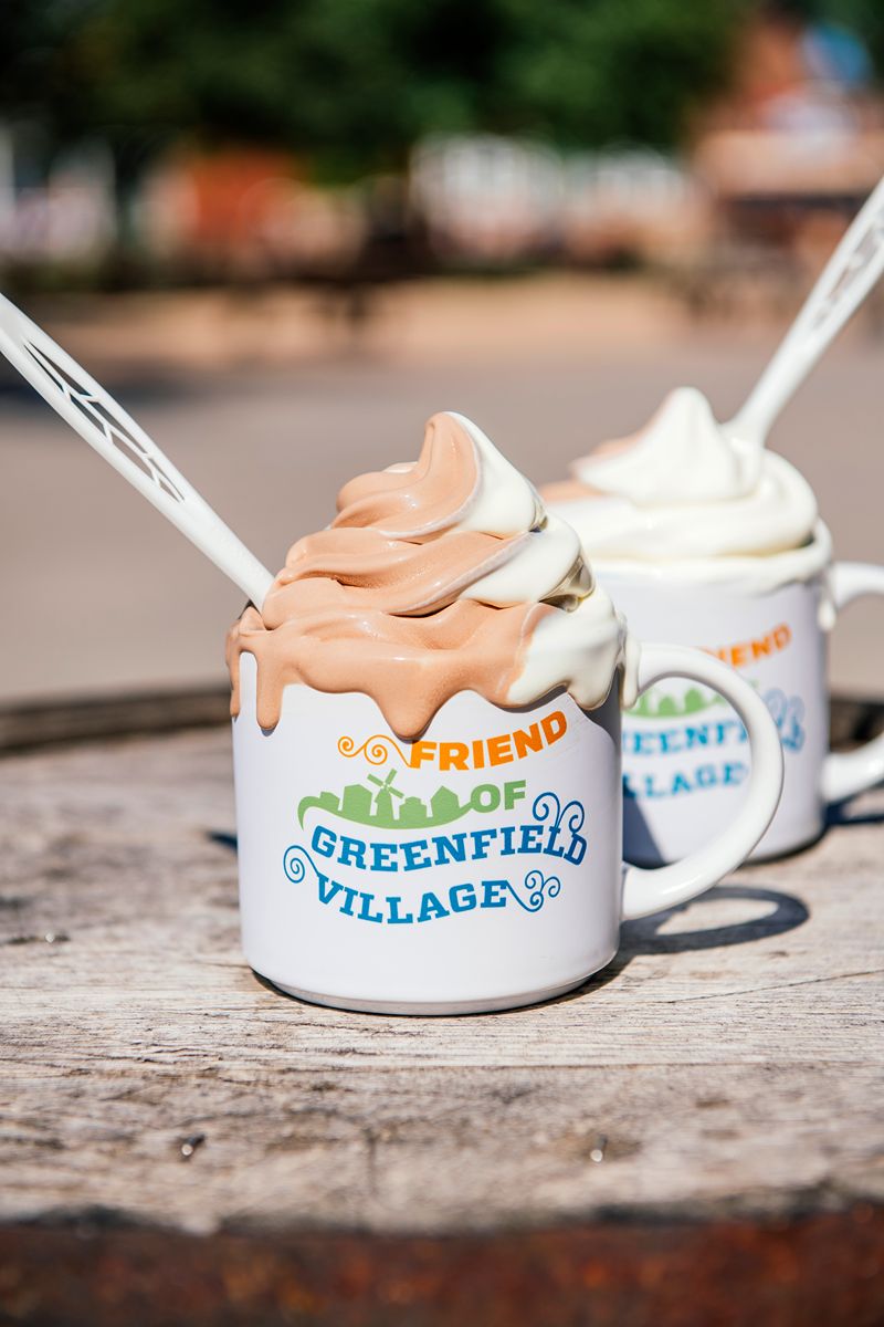 Two Friend of Greenfield Village Mugs hold swirled soft serve ice cream—one chocolate and vanilla, the other plain vanilla—each topped with a white plastic spoon. The mugs rest on a wooden surface outdoors in the center of the living history museum, surrounded by echoes of historic American artifacts.