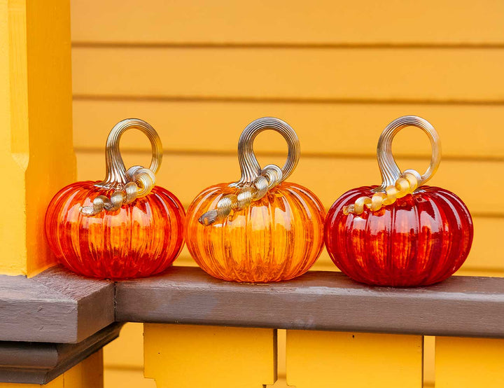 A Small Glass Pumpkin - Cherry sits on a gray ledge in front of a yellow wooden wall. This hand-blown glass pumpkin features a curly clear stem decorated with gold beads, making it the perfect collectible keepsake from Greenfield Village.