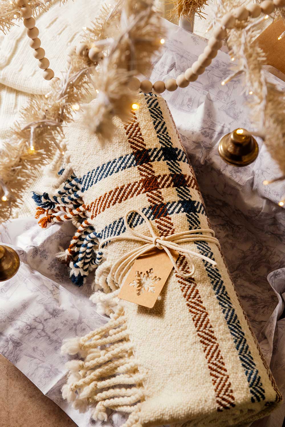 A Limited Edition Merino Wool Blanket, handwoven in Michigan with blue, brown, and orange stripes, comes tied with twine and a snowflake tag. It rests beneath a lit white Christmas tree decorated with ornaments.
