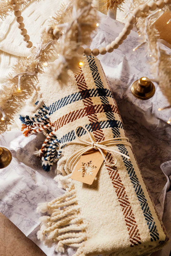 A Limited Edition Merino Wool Blanket, handwoven in Michigan with blue, brown, and orange stripes, comes tied with twine and a snowflake tag. It rests beneath a lit white Christmas tree decorated with ornaments.