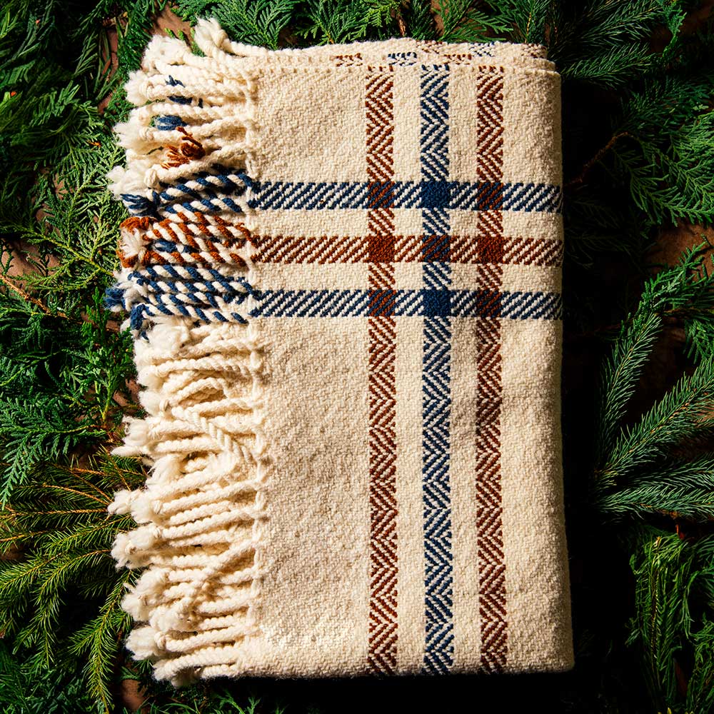 A folded Limited Edition Merino Wool Blanket in cream with blue and brown plaid stripes and fringe, surrounded by green pine branches.
