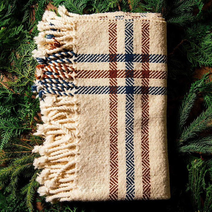 A folded Limited Edition Merino Wool Blanket in cream with blue and brown plaid stripes and fringe, surrounded by green pine branches.