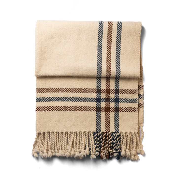 The Limited Edition Merino Wool Blanket, folded and featuring a beige base with black and brown plaid, is finished with twisted fringe tassels at the ends and displayed against a white background.