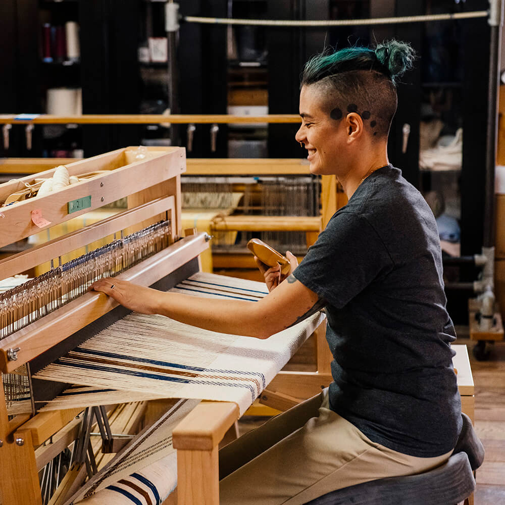 A person with short, partially shaved hair weaves a Limited Edition Merino Wool Blanket with blue and white stripes at a wooden loom, smiling and holding a weaving tool in a cozy indoor workspace.