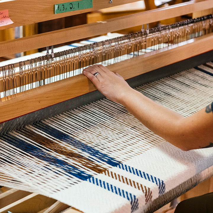 A hand adjusts threads on a wooden loom, weaving a Limited Edition Merino Wool Blanket in white with brown and blue accents; metal parts and the loom’s wooden frame are also visible.