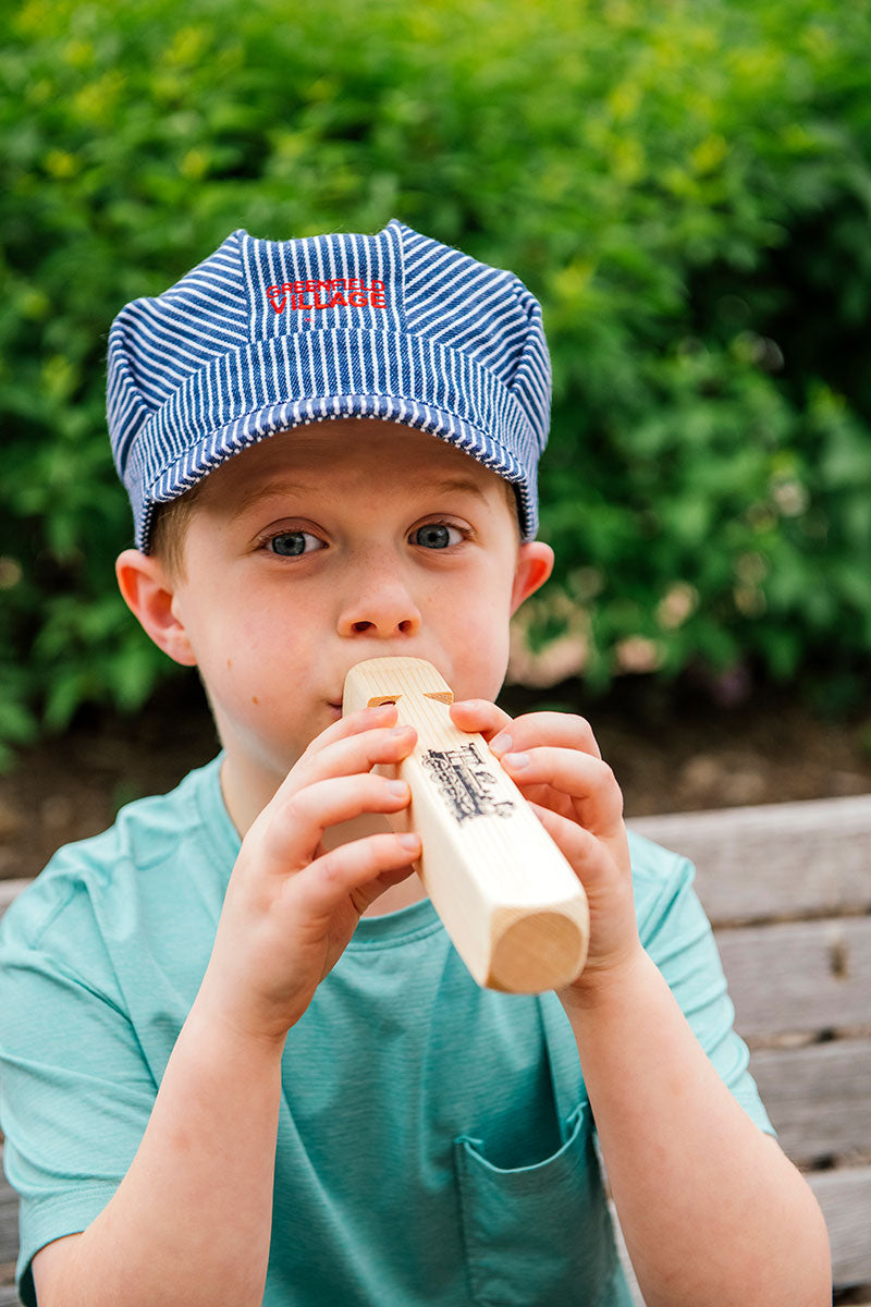 A young child wearing a Greenfield Village Youth Train Conductor Hat and a teal shirt plays a wooden train whistle while sitting on a bench outdoors, with greenery in the background.