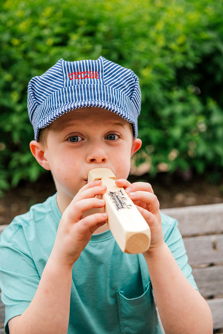 A young child wearing a Greenfield Village Youth Train Conductor Hat and a teal shirt plays a wooden train whistle while sitting on a bench outdoors, with greenery in the background.