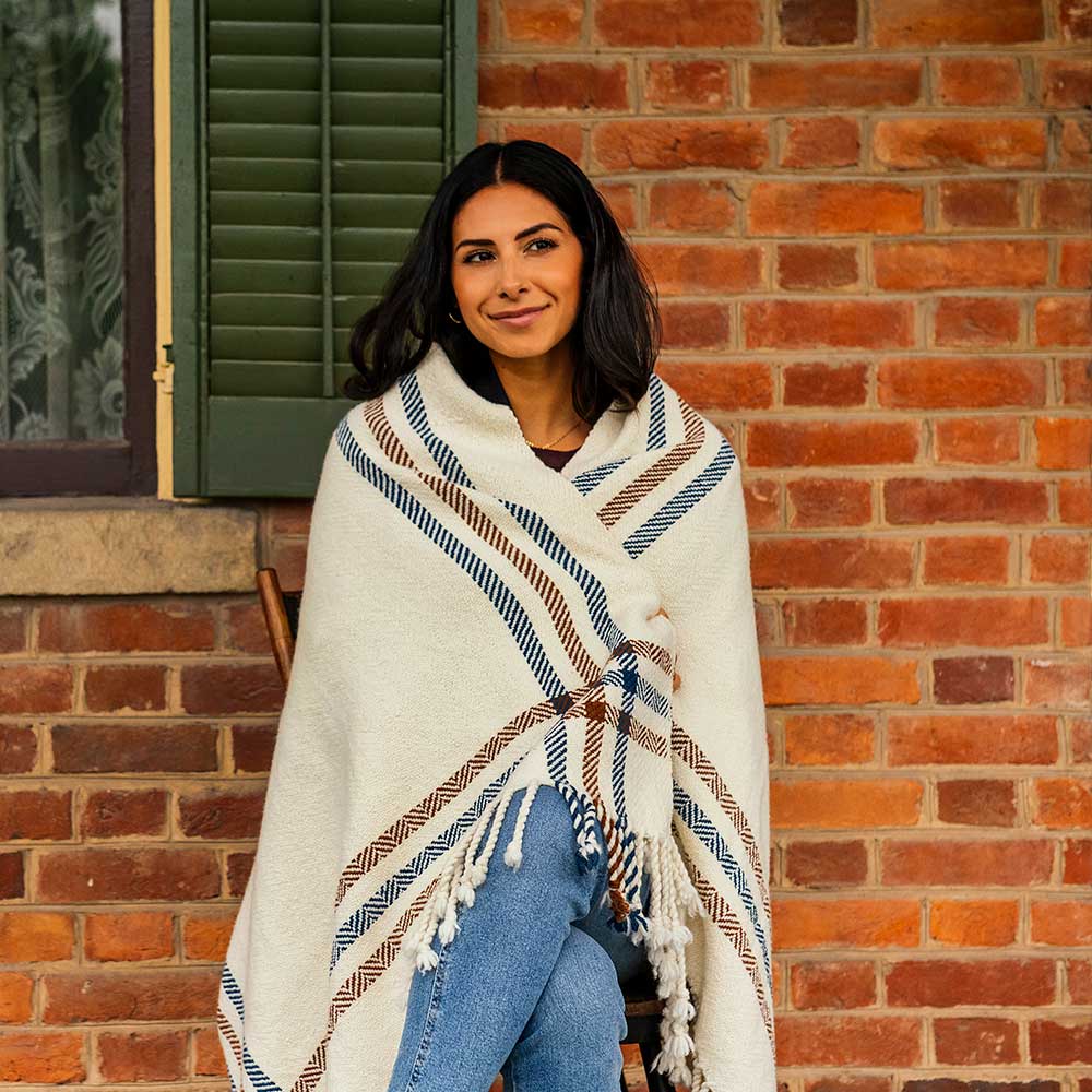 A woman with dark hair smiles softly at the camera, wrapped in a Limited Edition Merino Wool Blanket with brown and blue stripes, sitting on a wooden chair in front of a brick wall. She wears blue jeans. Made in Michigan.